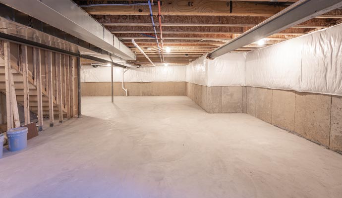 A spacious, clean, unfinished basement with concrete floors, exposed wooden wall frames and ceiling joists, and visible ductwork and plumbing.